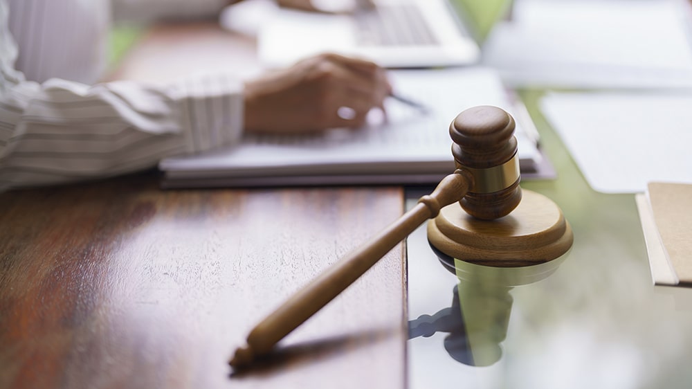 A gavel on a table with someone doing paperwork in the background.